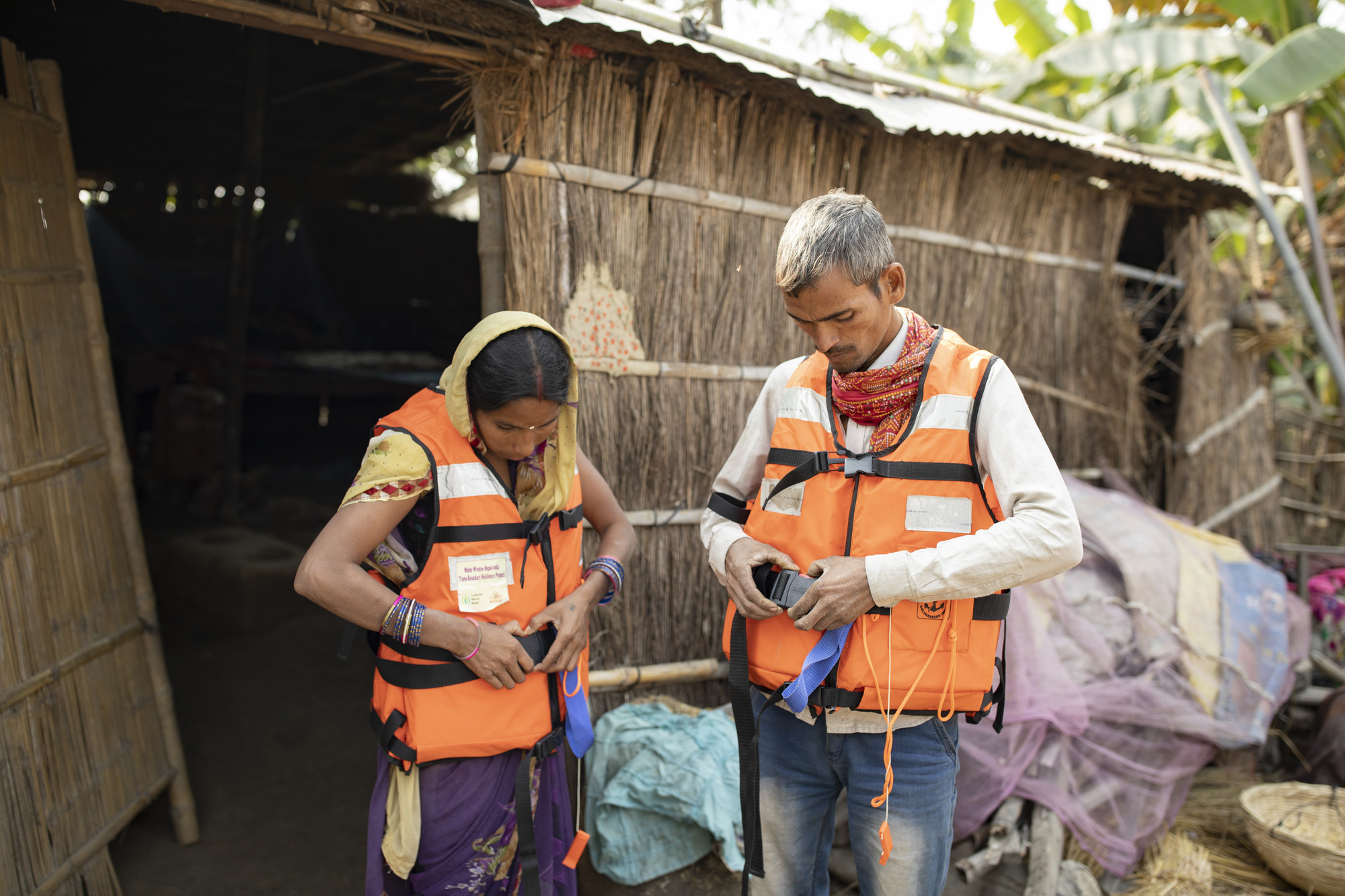 A woman and a man clip on orange life vests in front of a thatched house